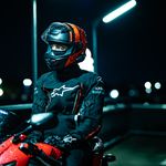 A motorcyclist in black and red gear sits on a parked bike under city lights at night.