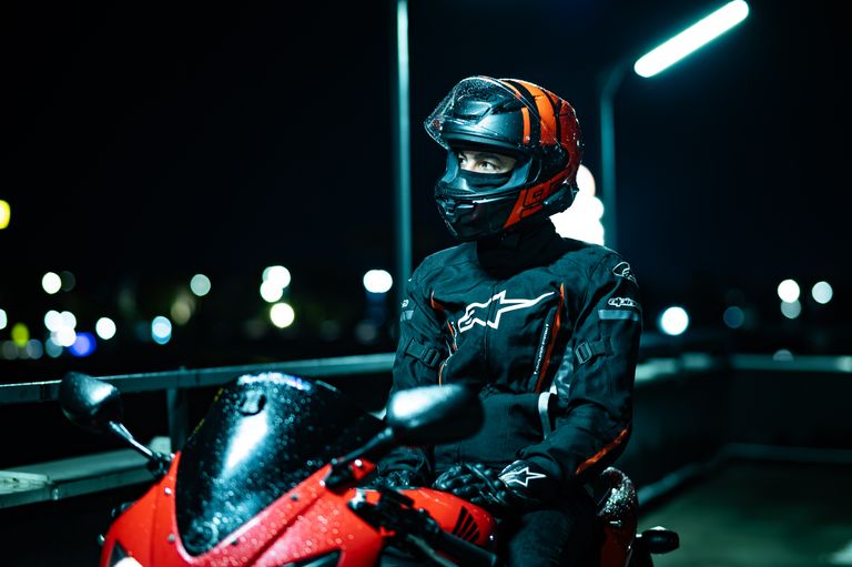 A motorcyclist in black and red gear sits on a parked bike under city lights at night.
