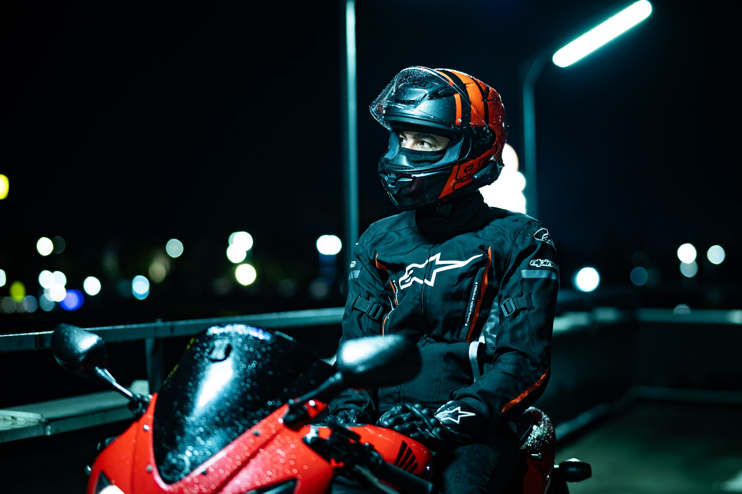 A motorcyclist in black and red gear sits on a parked bike under city lights at night.