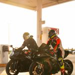 Two motorcyclists refueling at a gas station during sunset, in full racing gear.