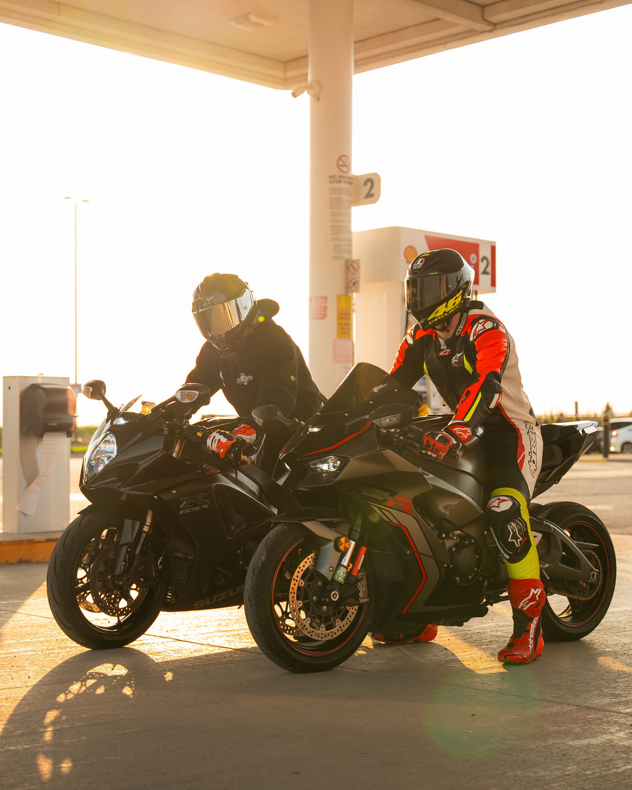 Two motorcyclists refueling at a gas station during sunset, in full racing gear.